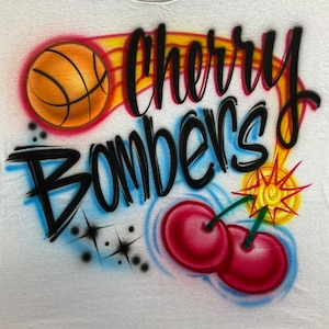 May include: A white t-shirt with a black and white airbrushed design featuring the words "Cherry Bombers" with a basketball and two cherries. The basketball is orange and has black lines. The cherries are red and have green stems. The background is white with blue and yellow accents.