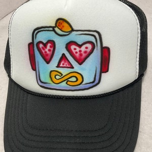May include: A white trucker hat with a black mesh back. The front of the hat features a hand-painted design of a blue robot face with red hearts for eyes, a yellow infinity symbol for a mouth, and a yellow antenna on top.