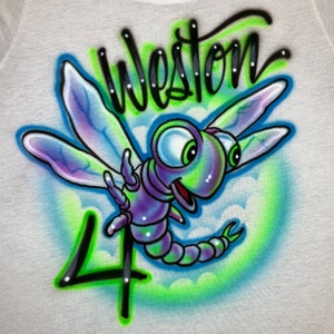Puede incluir: Una camiseta blanca con un diseño de libélula aerografiado en verde, azul y morado. La libélula tiene ojos grandes y una cara sonriente. El número "4" está aerografiado en negro debajo de la libélula. El nombre "Weston" está aerografiado en negro en la parte superior de la camiseta.