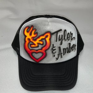 Può includere: Un cappello da camionista bianco e nero con una rete nera sul retro. La parte anteriore del cappello presenta una testa di cervo rossa, arancione e gialla con un cuore al centro. Il testo "Tyler & Amber" è scritto in corsivo nero sul lato destro del cappello.