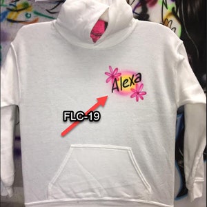 Puede incluir: Una sudadera con capucha blanca con un bolsillo delantero. La sudadera presenta el nombre "Alexa" en letras rosas y negras, acentuado con gr&aacute;ficos de flores rosas. El texto "FLC-19" tambi&eacute;n es visible.