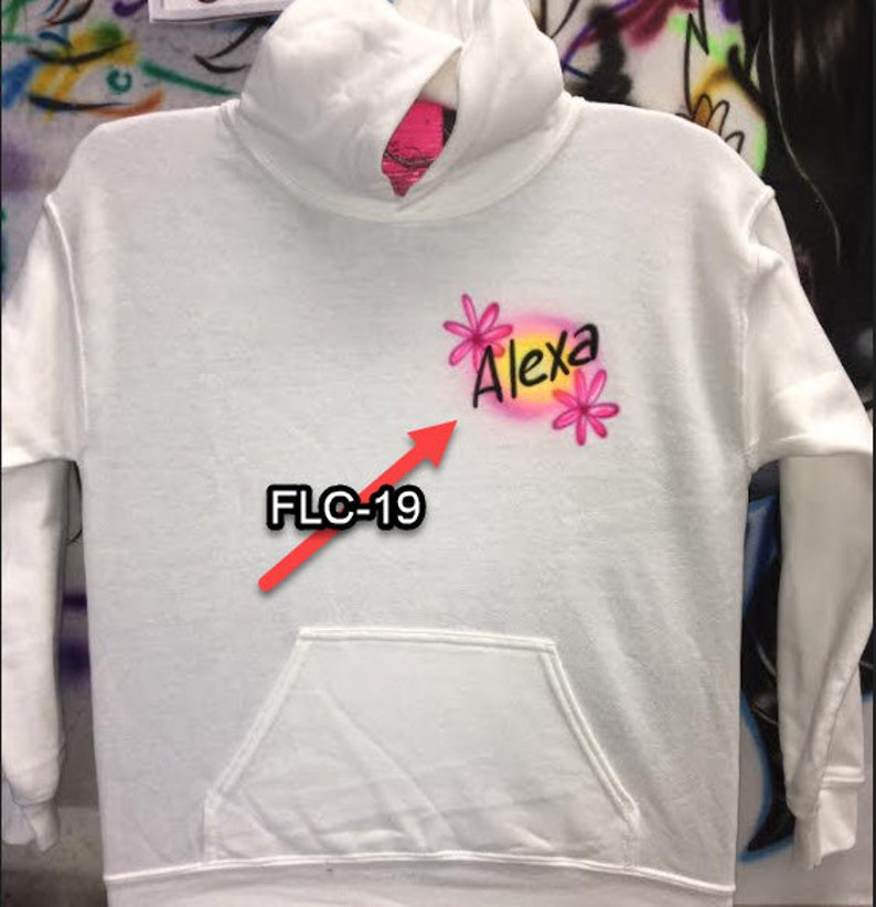 Puede incluir: Una sudadera con capucha blanca con un bolsillo delantero. La sudadera presenta el nombre "Alexa" en letras rosas y negras, acentuado con gr&aacute;ficos de flores rosas. El texto "FLC-19" tambi&eacute;n es visible.