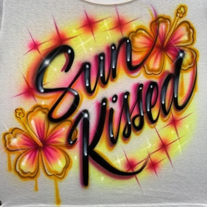 Pode incluir: Uma superfície de tecido branco com um design aerografado apresentando o texto cursivo preto Sun Kissed. O gráfico inclui duas grandes flores de hibisco laranja e amarelas e vários detalhes de estrelas rosa brilhante sobre um fundo amarelo.
