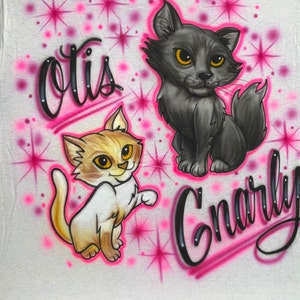 May include: White t-shirt with a pink and black airbrushed design featuring two cartoon cats. The cats have yellow eyes and are surrounded by pink stars. The text "Otis" and "Gnarly" are written in black cursive.
