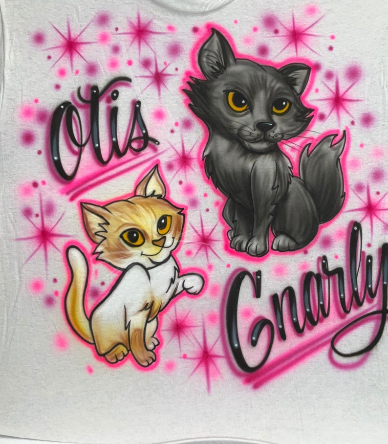 May include: White t-shirt with a pink and black airbrushed design featuring two cartoon cats. The cats have yellow eyes and are surrounded by pink stars. The text "Otis" and "Gnarly" are written in black cursive.