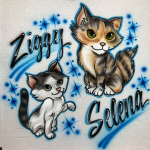 May include: A white t-shirt with a blue and white airbrushed design of two cartoon cats. The cat on the left is white with black and gray markings and is named "Ziggy". The cat on the right is brown, black, and white and is named "Selena".