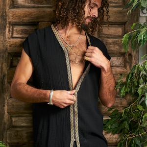 Pode incluir: Uma camisa preta sem mangas com uma guarni&ccedil;&atilde;o dourada na frente. A camisa est&aacute; aberta na frente, revelando um colar de corrente prateada com um pingente dourado.
