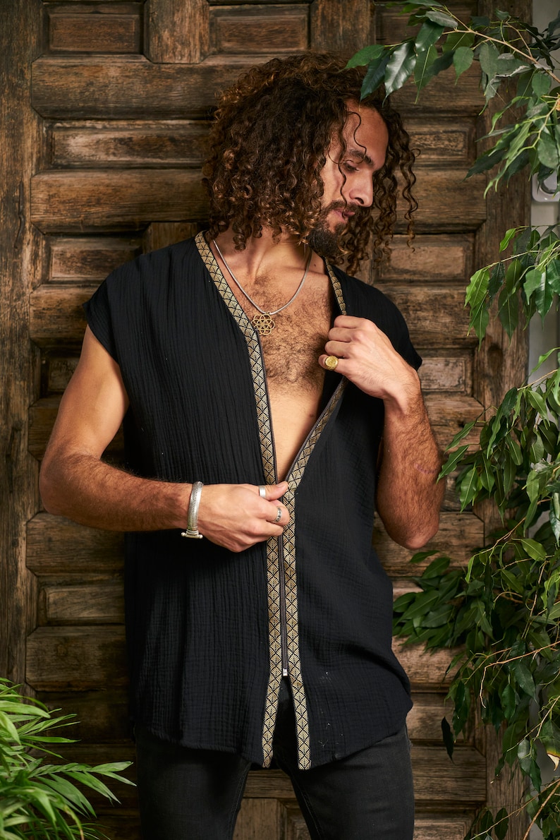 Pode incluir: Uma camisa preta sem mangas com uma guarni&ccedil;&atilde;o dourada na frente. A camisa est&aacute; aberta na frente, revelando um colar de corrente prateada com um pingente dourado.