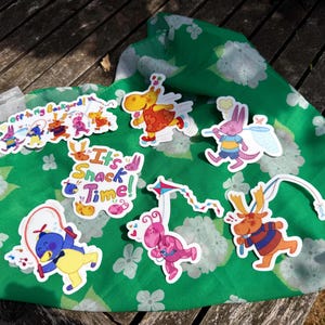 May include: A selection of vibrant stickers depicting cartoon characters, arranged on a green floral fabric. The stickers showcase characters in playful poses, with text reading "Off to my Backyard!" and "It's Snack Time!"