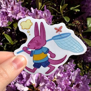 May include: A die-cut sticker featuring a cartoon kangaroo in a blue and yellow striped shirt and blue shorts, carrying a butterfly net. A thought bubble and a butterfly are also depicted. The sticker is held in front of a background of purple flowers.