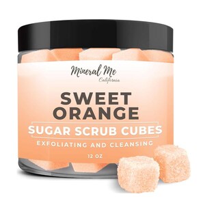 May include: A clear jar filled with orange sugar scrub cubes. The jar has a black lid and a white label that reads "Mineral Me California Sweet Orange Sugar Scrub Cubes Exfoliating and Cleansing 12 oz."