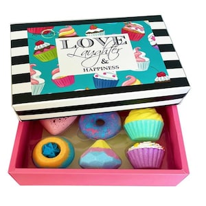 May include: A pink box with a black and white striped lid. The lid has a design of cupcakes with the text "Love Laughter & Happiness". The box contains 8 colorful bath bombs shaped like cupcakes and donuts.