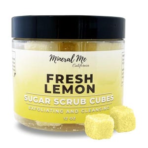 May include: A clear jar with a black lid filled with yellow sugar scrub cubes. The jar has a label that reads "Mineral Me California Fresh Lemon Sugar Scrub Cubes Exfoliating and Cleansing 12 oz". Two yellow sugar scrub cubes are in the foreground.