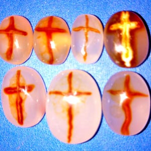 May include: Seven oval-shaped, polished stones with a cross design. The crosses are reddish-brown against a translucent, off-white background. The stones vary in colour and pattern, with some having more pronounced brown hues.