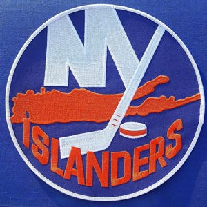 Peut inclure: Un écusson brodé circulaire sur fond bleu avec les lettres blanches NY, une silhouette de carte orange, une crosse de hockey blanche, un palet de hockey blanc à rayures orange et le texte orange ISLANDERS avec une bordure blanche.
