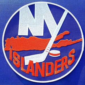 May include: A circular embroidered patch featuring a blue background with the white letters NY, an orange map silhouette, a white hockey stick, a white hockey puck, and the orange text ISLANDERS, all bordered by a white stitched edge.