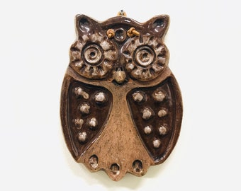 Könnte beinhalten: Ein brauner Keramik-Eulen-Wandbehang. Die Eule hat große, detaillierte Augen, einen strukturierten Körper und eine kleine Schlaufe zum Aufhängen. Der Körper der Eule ist hellbraun mit dunkelbraunen Akzenten und weißen Punkten.