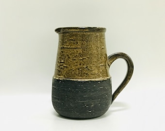 May include: A small, brown and black ceramic pitcher with a handle. The pitcher has a textured surface and a wide, round base.