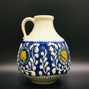 Puede incluir: Una jarra de cerámica blanca con un diseño floral azul y amarillo. La jarra tiene un cuerpo redondo y un asa.