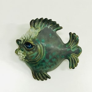 Puede incluir: Una escultura de pescado de cerámica con un esmalte verde y azul. El pez tiene un cuerpo grande y redondo y una cola pequeña y puntiaguda. Los ojos del pez son azules y la boca del pez está abierta.