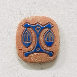Könnte beinhalten: Eine quadratische Keramikfliese in Terrakotta-Farbe mit einem blauen Waage-Symbol. Das Symbol zeigt eine stilisierte Waage mit zwei hängenden Schalen. Die Fliese hat eine strukturierte Oberfläche und eine kleine "LB"-Initiale.