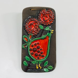 May include: A ceramic wall hanging with a green bird with red and orange spots, surrounded by green leaves and red and orange flowers.
