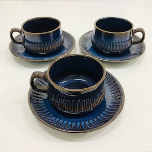 May include: Three blue ceramic teacups and saucers with a brown glaze and a textured design. The cups and saucers are arranged in a row on a white surface.