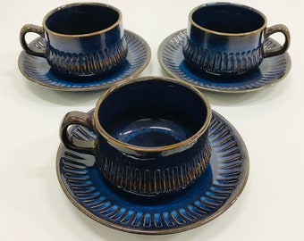 Puede incluir: Tres tazas de té y platillos de cerámica azul con un esmalte marrón y un diseño texturizado. Las tazas y los platillos están dispuestos en una fila sobre una superficie blanca.