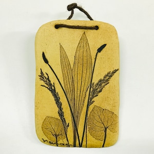 May include: A beige ceramic wall hanging with a pressed leaf design. The leaves are brown and black and are arranged in a cluster. The wall hanging has a brown string attached to the top for hanging.