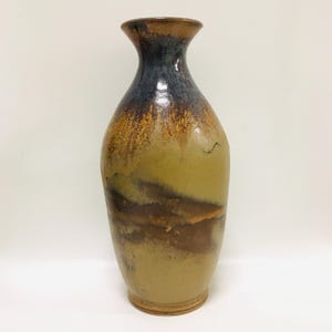 May include: A tall, handcrafted ceramic vase with a slender neck and a wide, rounded body. The vase features a gradient of colours, transitioning from a dark blue-brown at the top to earthy tones of brown, tan, and olive green. The vase is approximately 30 cm tall.