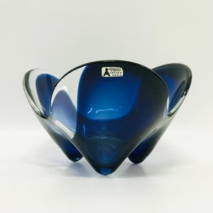 May include: A blue glass bowl with a unique, flower-like shape. The bowl has three legs and a label that reads "Sweden Kosta Boda".