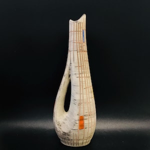 Può includere: Un vaso in ceramica bianca con un design geometrico in arancione e blu. Il vaso ha una forma unica e curva con un manico tagliato.