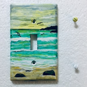May include: A light switch cover painted with a seascape scene. The cover features a blue and green ocean with white waves, a sandy beach, and a yellow sky with birds flying overhead.