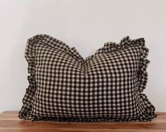 Könnte beinhalten: Ein rechteckiges Kissen mit einem schwarz-beigen Vichy-Muster. Das Kissen hat einen gerüschten Rand und liegt auf einer Holzoberfläche. Das Kissen ist ein dekoratives Element für ein Bett oder Sofa.