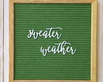 May include: A green felt letter board with a wooden frame. The board has white letters spelling out "sweater weather".