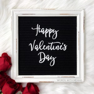 May include: White framed letter board with black felt background and white letters spelling "Happy Valentine's Day". The board is surrounded by red roses on a white fuzzy surface.