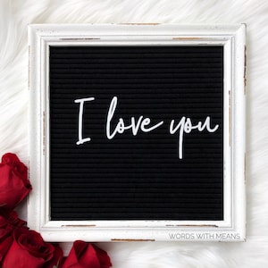 May include: A white framed letter board with a black felt background. The board has the words "I love you" written on it in white letters. The board is sitting on a white fluffy surface with red roses in the foreground.