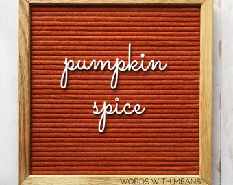 May include: A brown felt letter board with a wooden frame displays the text "pumpkin spice" in white letters. The board has a white background and is sitting on a white wooden surface. The text "WORDS WITH MEANS" is printed in white at the bottom of the board.