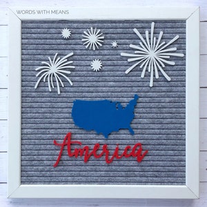 Puede incluir: Un tablero de letras de fieltro gris con un contorno azul de los Estados Unidos y la palabra "America" en letras rojas. Recortes de papel blanco de fuegos artificiales est&aacute;n dispuestos alrededor del mapa.