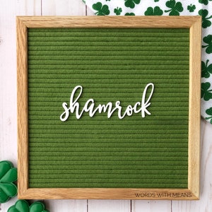 May include: A green felt letter board with a white wooden cutout of the word "shamrock" on it. The board is framed in wood and has a white background.