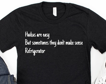 Puede incluir: Camiseta negra con texto blanco que dice "Haikus are easy But sometimes they don't make sense Refrigerator". La camiseta está colocada sobre una superficie blanca con un par de zapatillas blancas, unos vaqueros azules y unos auriculares negros.
