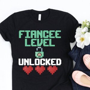 May include: Black t-shirt with the text "FIANC&Eacute;E LEVEL UNLOCKED" in pixelated green and white font. The design includes a pixelated lock with a smiley face and three red hearts. The shirt is paired with dark blue jeans and pink ballet flats.