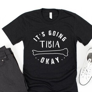 May include: Black t-shirt with white text that reads "It's going tibia okay" with a white outline of a bone.