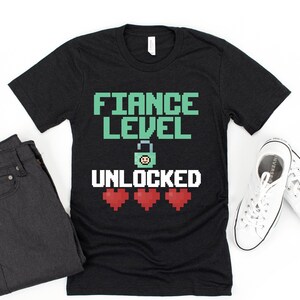 May include: Black t-shirt with the text "FIANCE LEVEL UNLOCKED" in a pixelated font. The text is green and white. There are three red hearts below the text.
