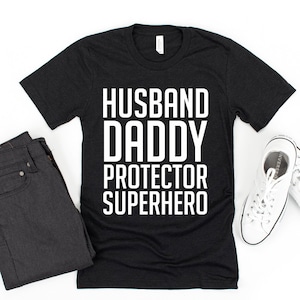 May include: Black t-shirt with white text that reads "Husband Daddy Protector Superhero".