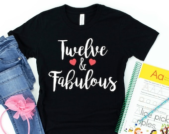 Puede incluir: Camiseta negra con el texto "Twelve & Fabulous" en letras blancas con dos corazones rojos. La camiseta está colocada sobre un fondo blanco con un par de pantalones vaqueros azules, una diadema rosa y un libro de trabajo.