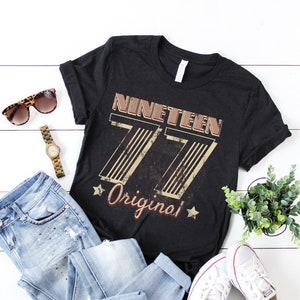 May include: Black t-shirt with a vintage-style graphic that says "Nineteen 77 Original" in white and red lettering.