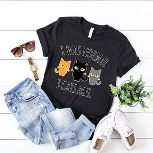 May include: Black t-shirt with a white graphic that says "I was normal 3 cats ago." The graphic features three cartoon cats, one orange, one black, and one gray.