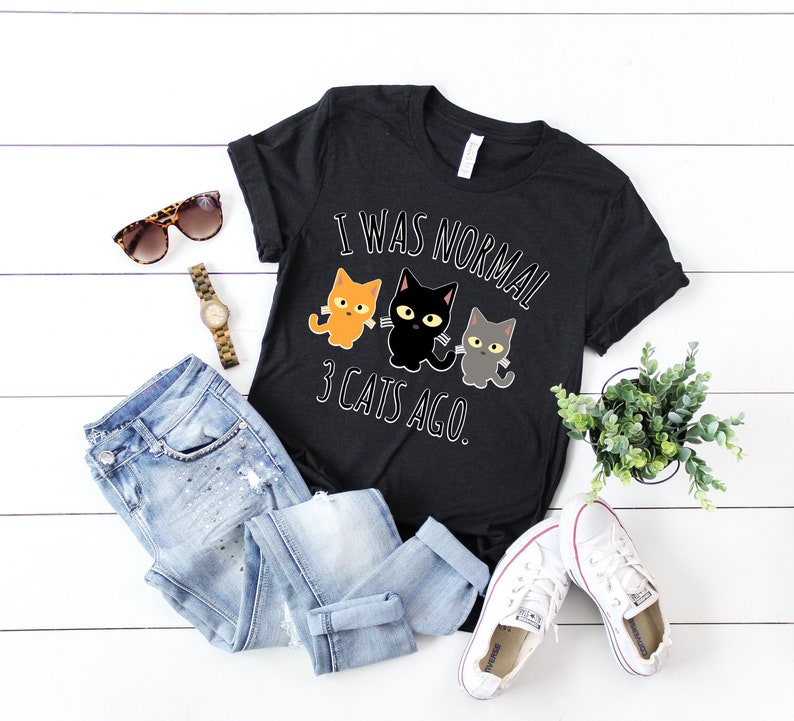 May include: Black t-shirt with a white graphic that says "I was normal 3 cats ago." The graphic features three cartoon cats, one orange, one black, and one gray.