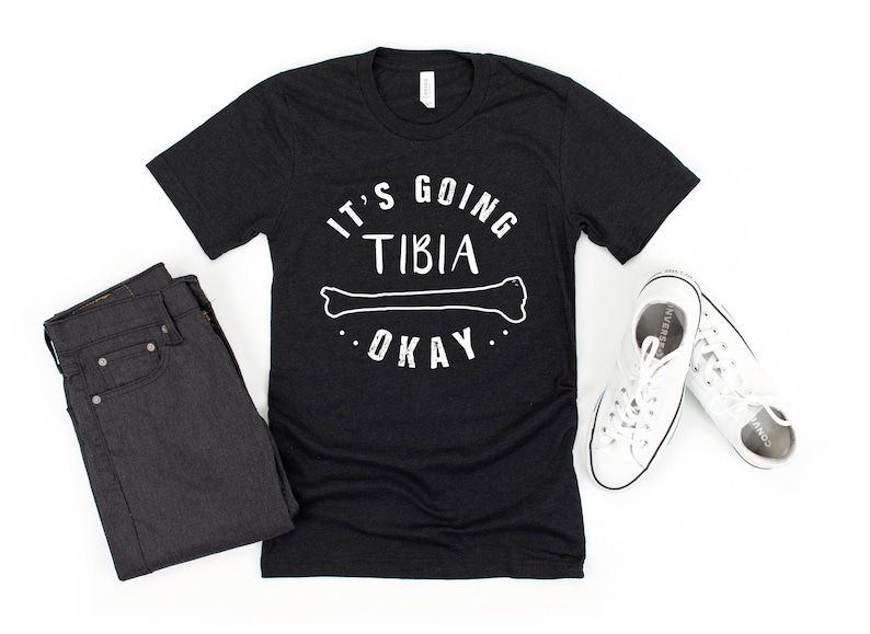 May include: Black t-shirt with white text that reads "It's going tibia okay" with a white outline of a bone.